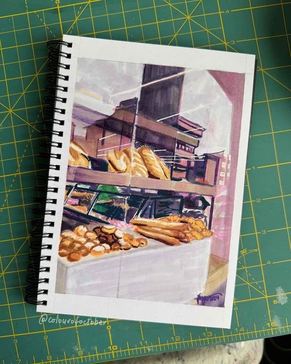 Sketchbook open to a page with a marker drawing of a bakery, on a green cutting mat