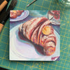 A colorful, square, textured oil painting of a croissant on a plate, sits on a green cutting mat next to paintbrushes.