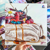 Hand holding a fine art print of a pavlova dessert topped with berries, cream, and syrup, presented in a colorful and realistic style.