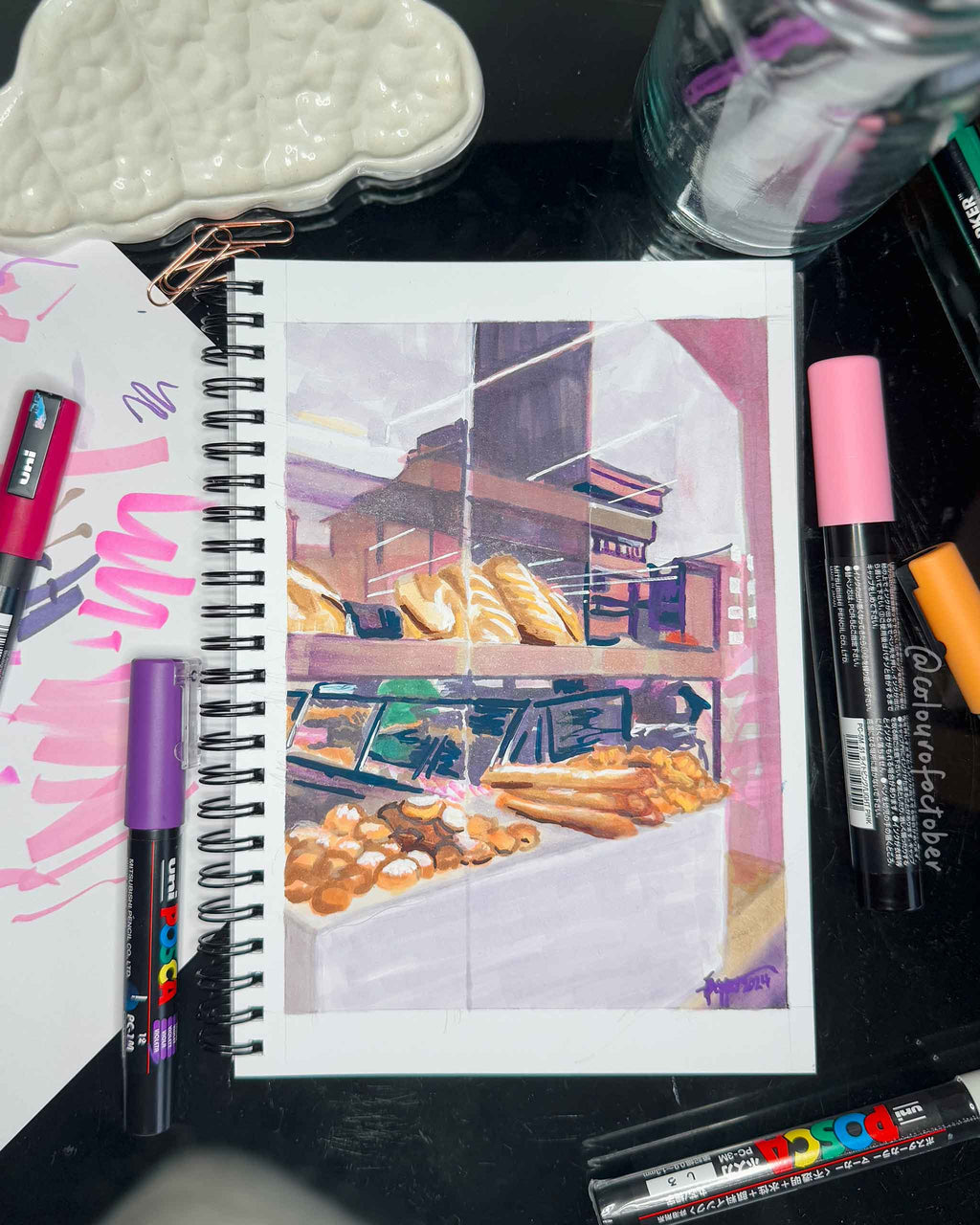 Artist's sketchbook with a drawing of a bakery on a desk with markers around