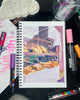 Artist's sketchbook with a drawing of a bakery on a desk with markers around
