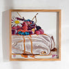 A colourful fine art print of a digital painting featuring a pavlova dessert topped with a white and yellow flower, berries, cream, and syrup, in a oak coloured frame, on a bright white wall.