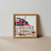 A colourful fine art print of a digital painting featuring a pavlova dessert topped with a white and yellow flower, berries, cream, and syrup, in a light brown frame, against a neutral background. 